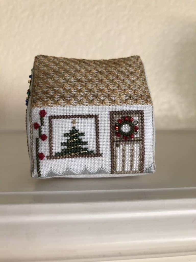 A cross-stitched Christmas ornament in the shape of a house with a Christmas tree in the window and a wreath on the door